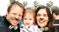 Parents with cat whisker face paint hold young child during Purim celebration